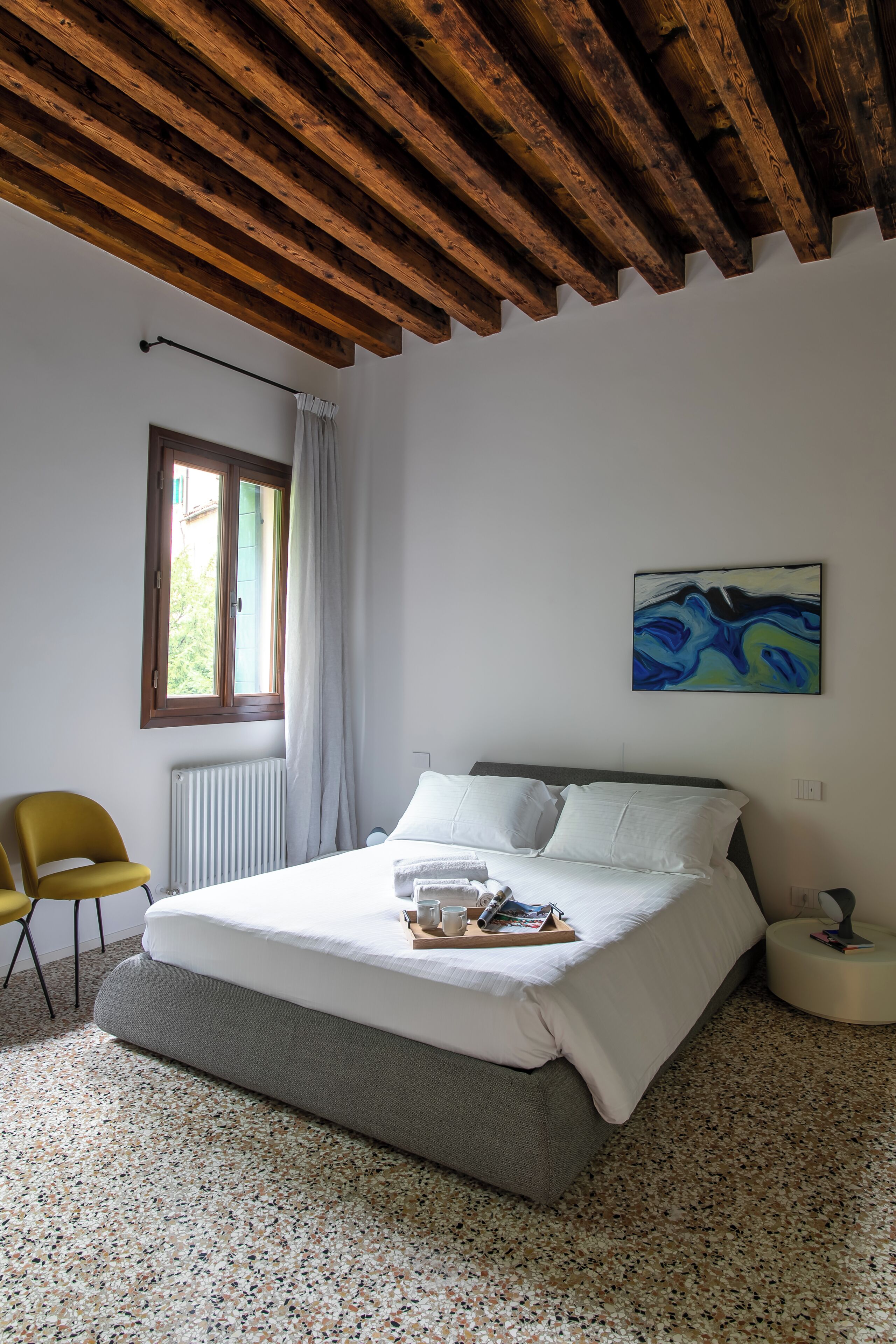 Een slaapkamer met een bed, een raam met gordijnen, een radiator, een stoel en een schilderij aan de muur.