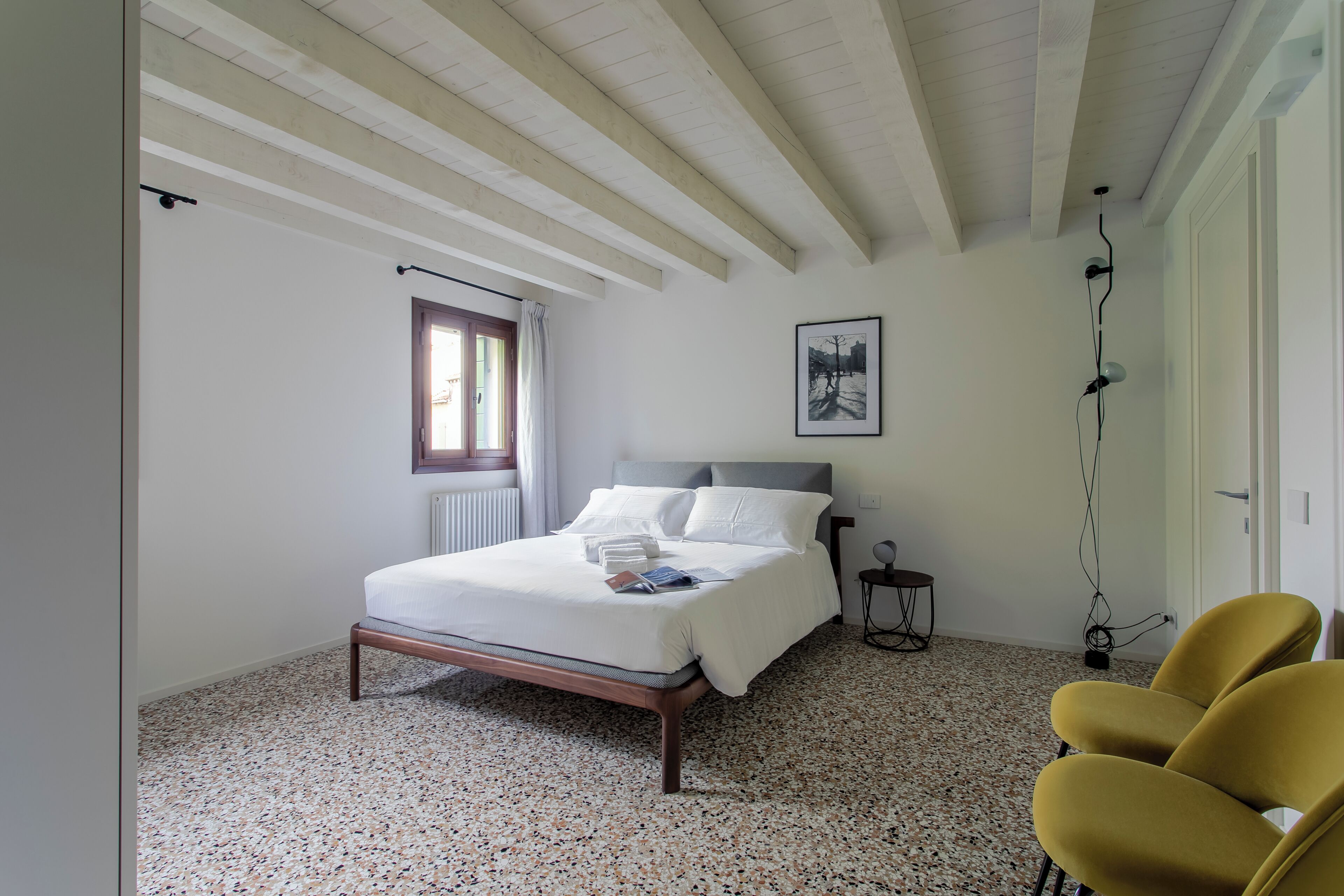 A bedroom with a bed, a window, a radiator, a framed picture, and a chair.