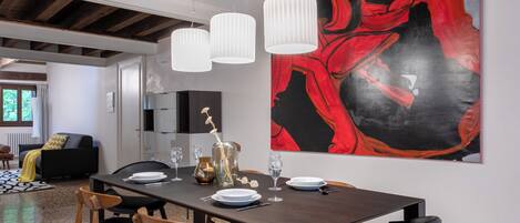 Comedor con mesa de madera dispuesta para cuatro, lámparas colgantes blancas y un cuadro abstracto grande en la pared.