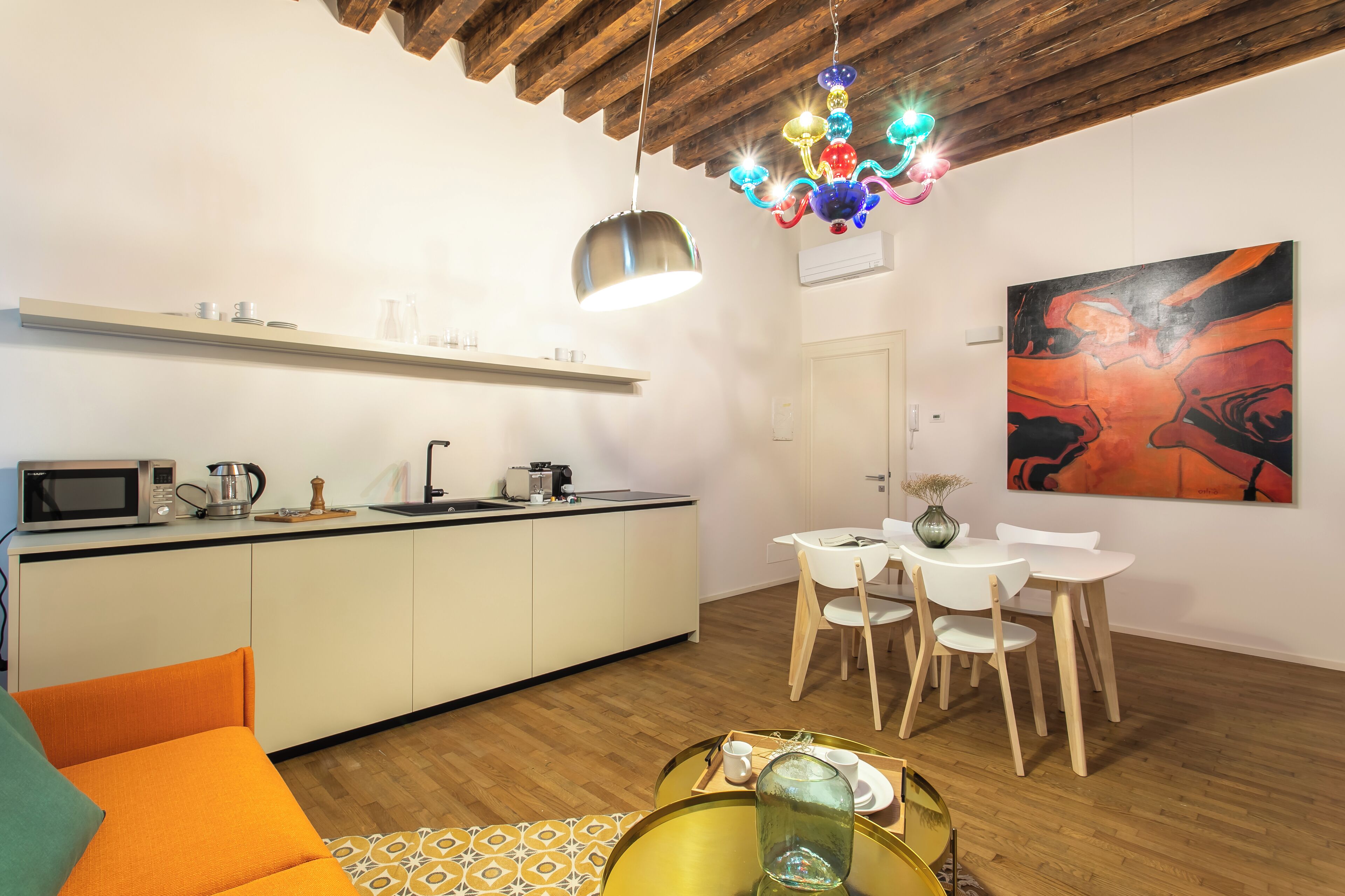 A modern kitchen with a microwave, kettle, and coffee maker. There is a dining area with a white table and chairs. A colorful chandelier hangs from the ceiling.