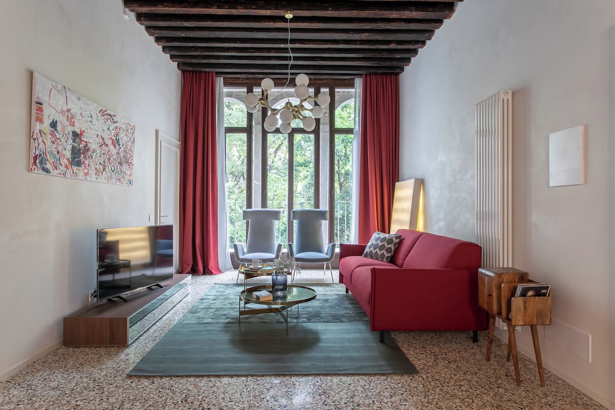 A modern living room with a red sofa, two armchairs, a coffee table, a TV, and a large window with curtains.