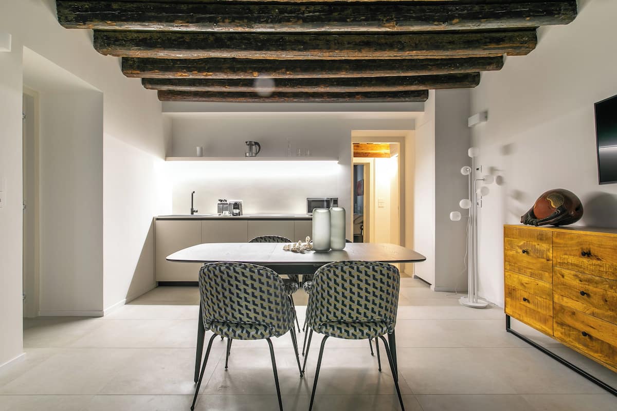 A modern kitchen with a central island, two patterned chairs, a wooden cabinet, and a flat-screen TV on the wall.
