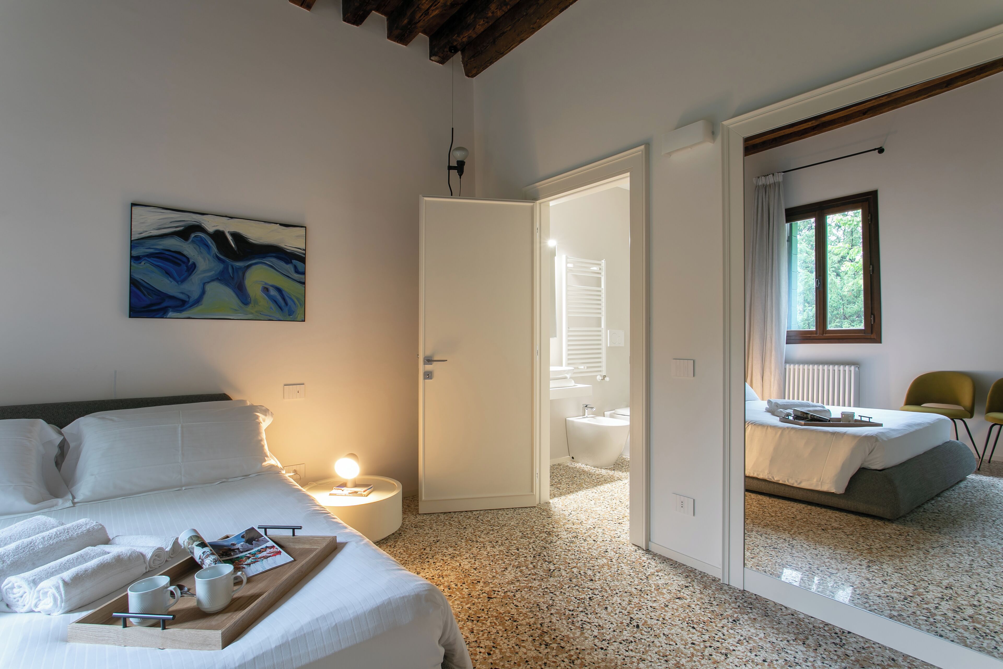 A hotel room with a bed, a bedside table with a lamp, a painting on the wall, and a bathroom visible through an open door.