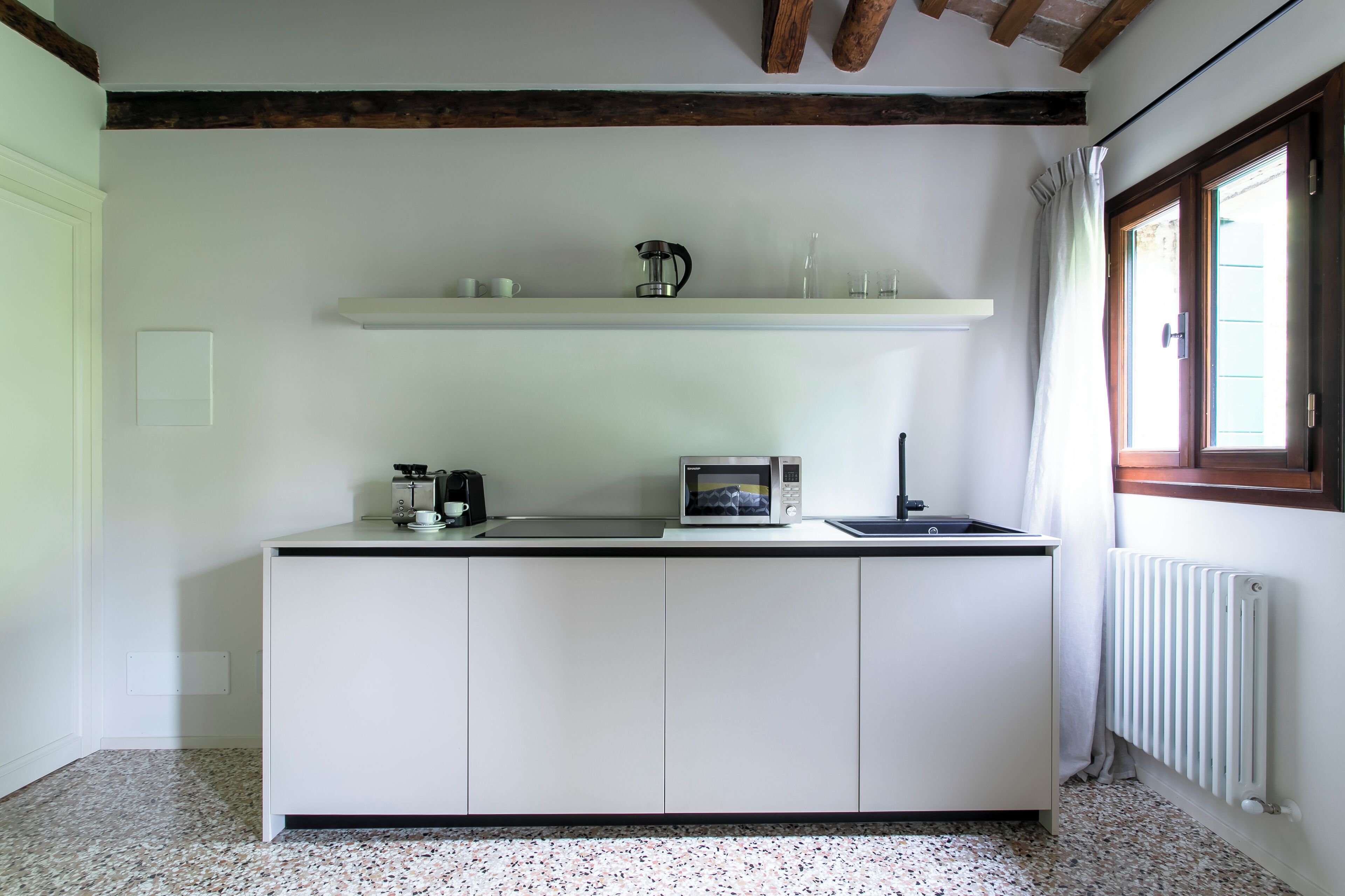 A compact kitchen with white cabinets, a microwave, a kettle, and a small sink. There is a window with curtains and a radiator on the wall.