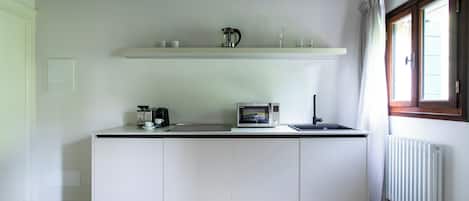 A compact kitchen with white cabinets, a microwave, a kettle, and a small sink. There is a window with curtains and a radiator on the wall.
