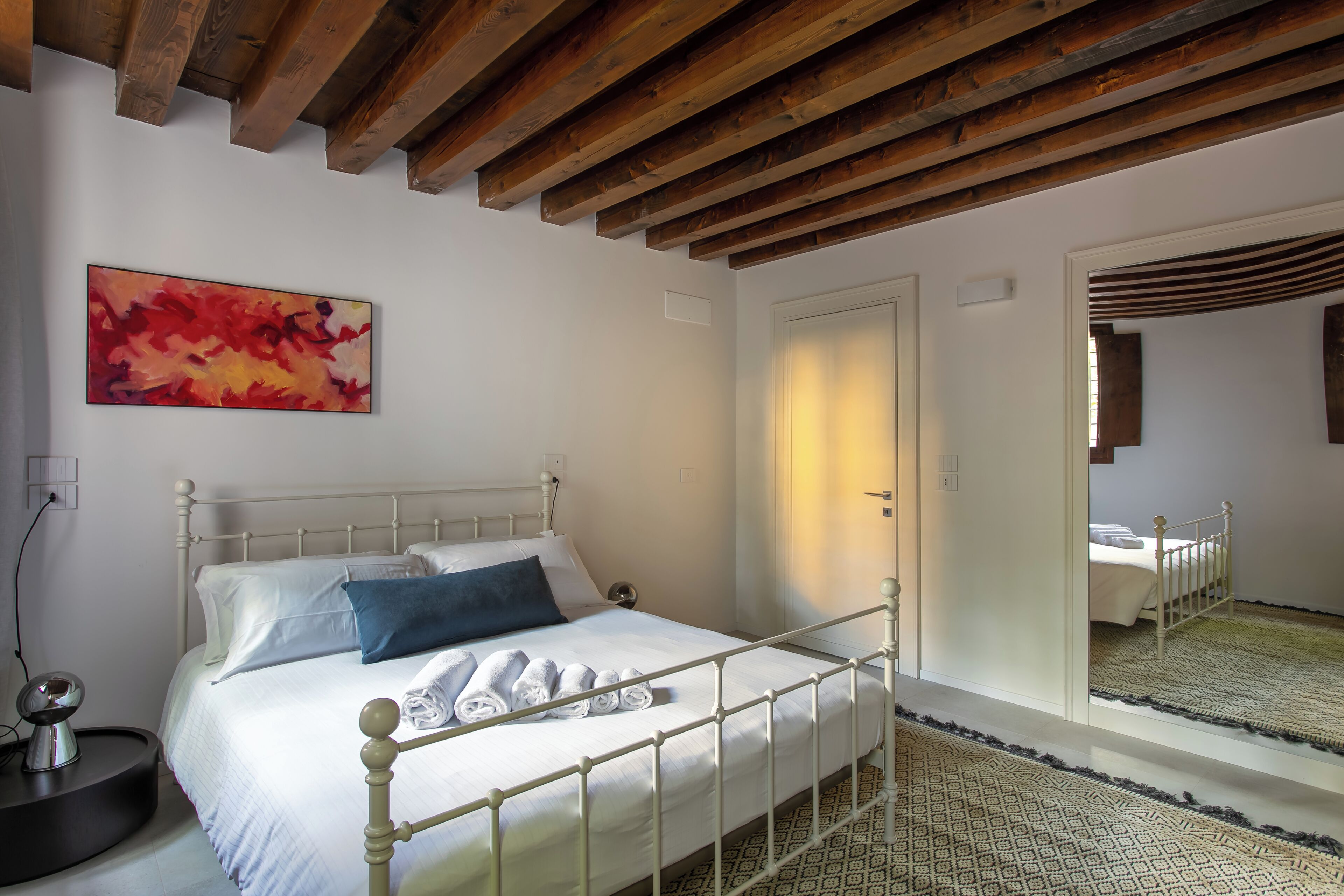 A bedroom with a bed, a bedside table, a mirror, and a wooden ceiling.