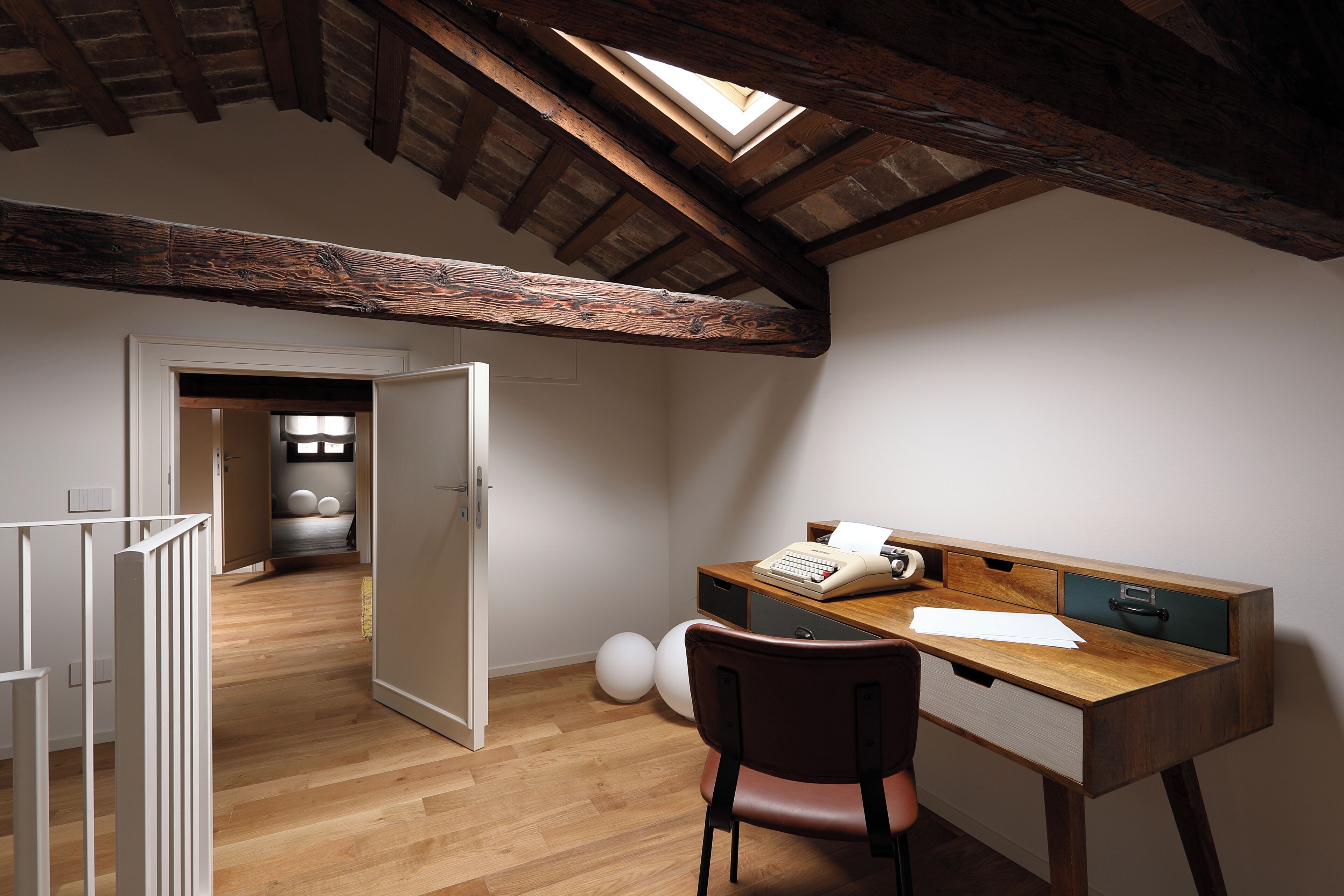 Una habitación con escritorio de madera, una silla, un tragaluz y una puerta que da a otra estancia.