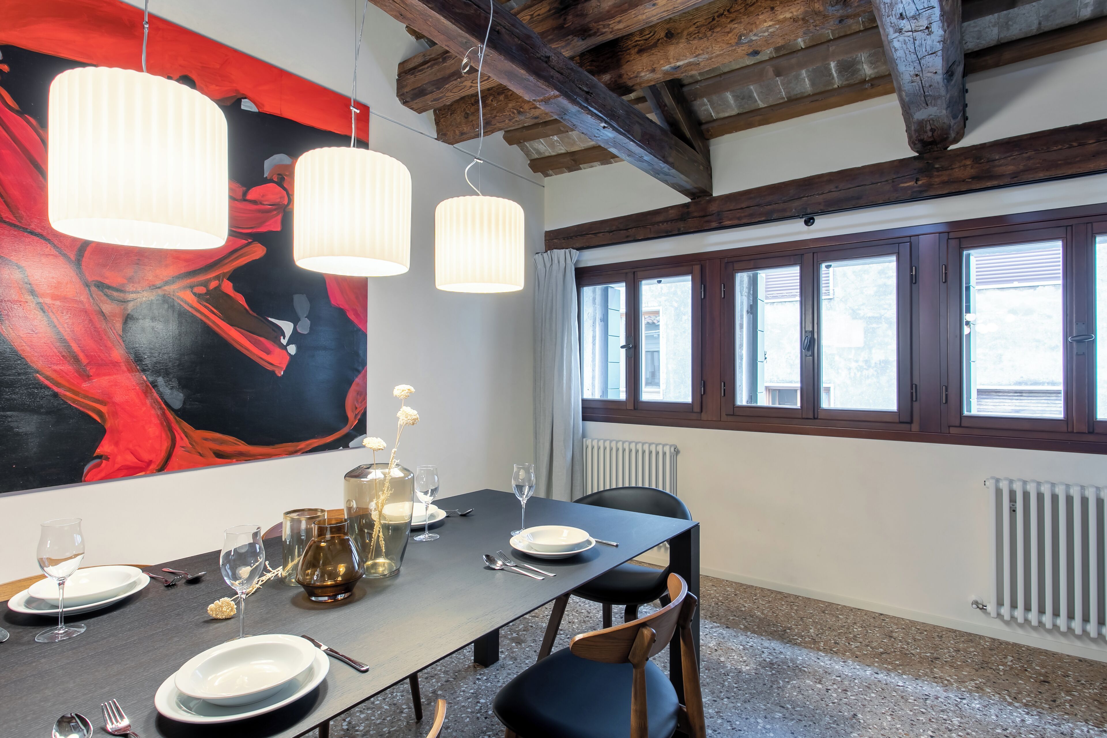 Un comedor con una mesa dispuesta para cuatro personas, que incluye un cuadro abstracto de gran tamaño, lámparas colgantes y vistas al exterior.