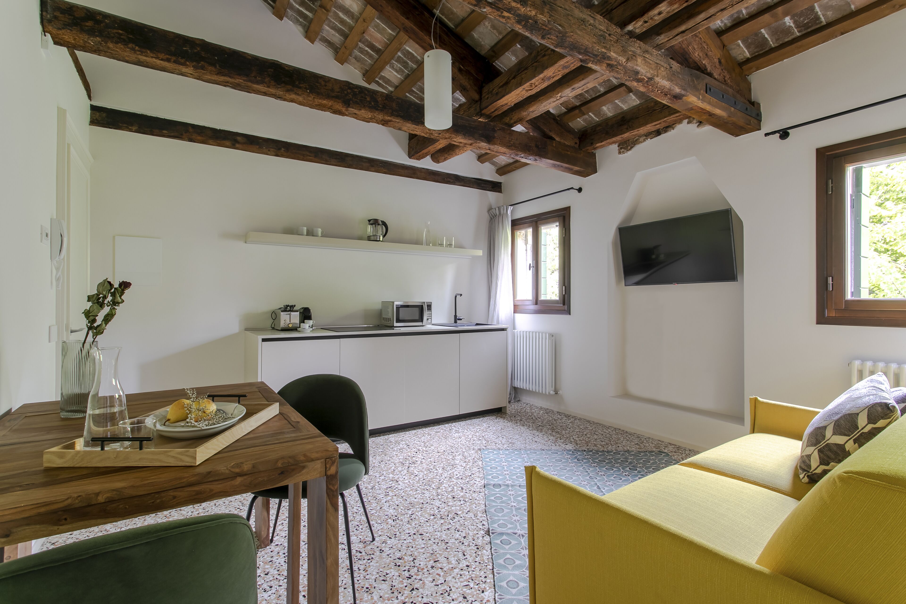 Una habitación acogedora con una mesa de comedor de madera, una silla verde, un sofá amarillo y una zona de cocina con microondas y estantería.