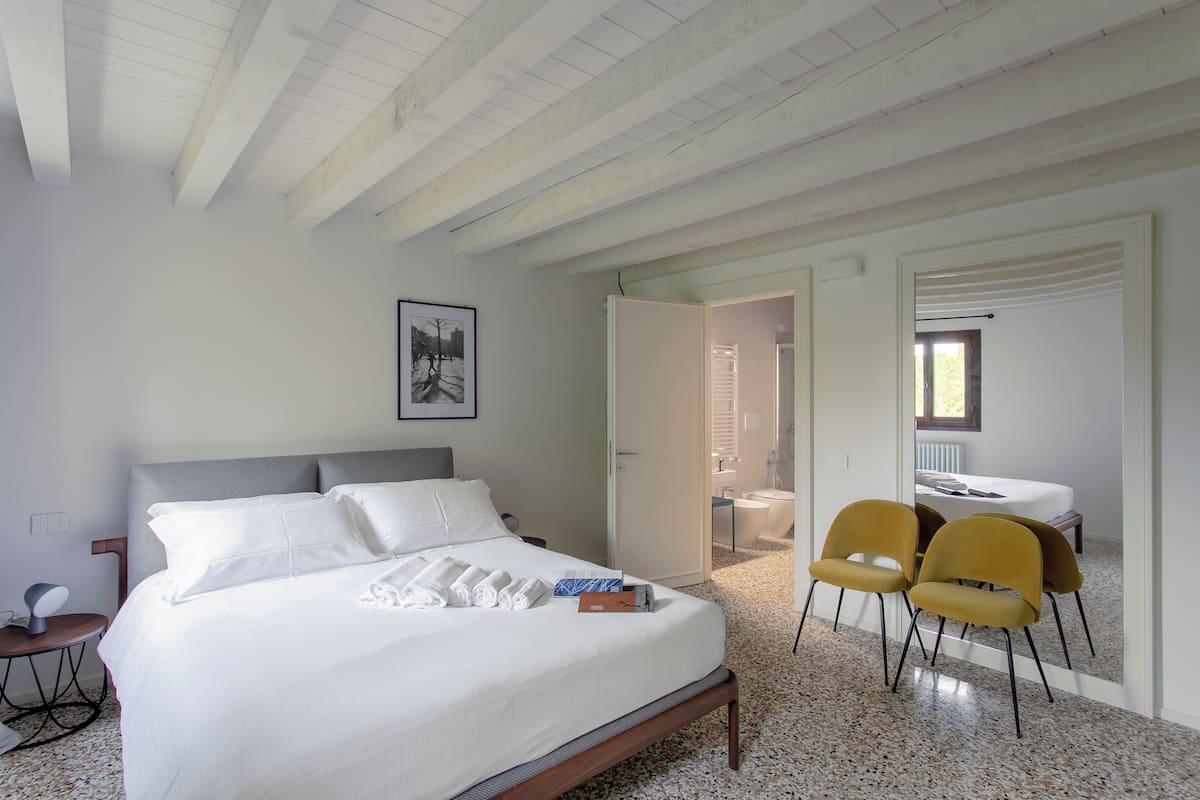 A bedroom with a bed, a bedside table, a chair, and a bathroom visible through an open door.