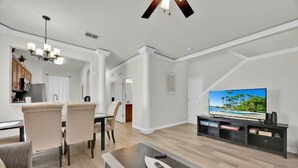 A modern living room with a dining area, a kitchen, and a TV displaying a beach scene.