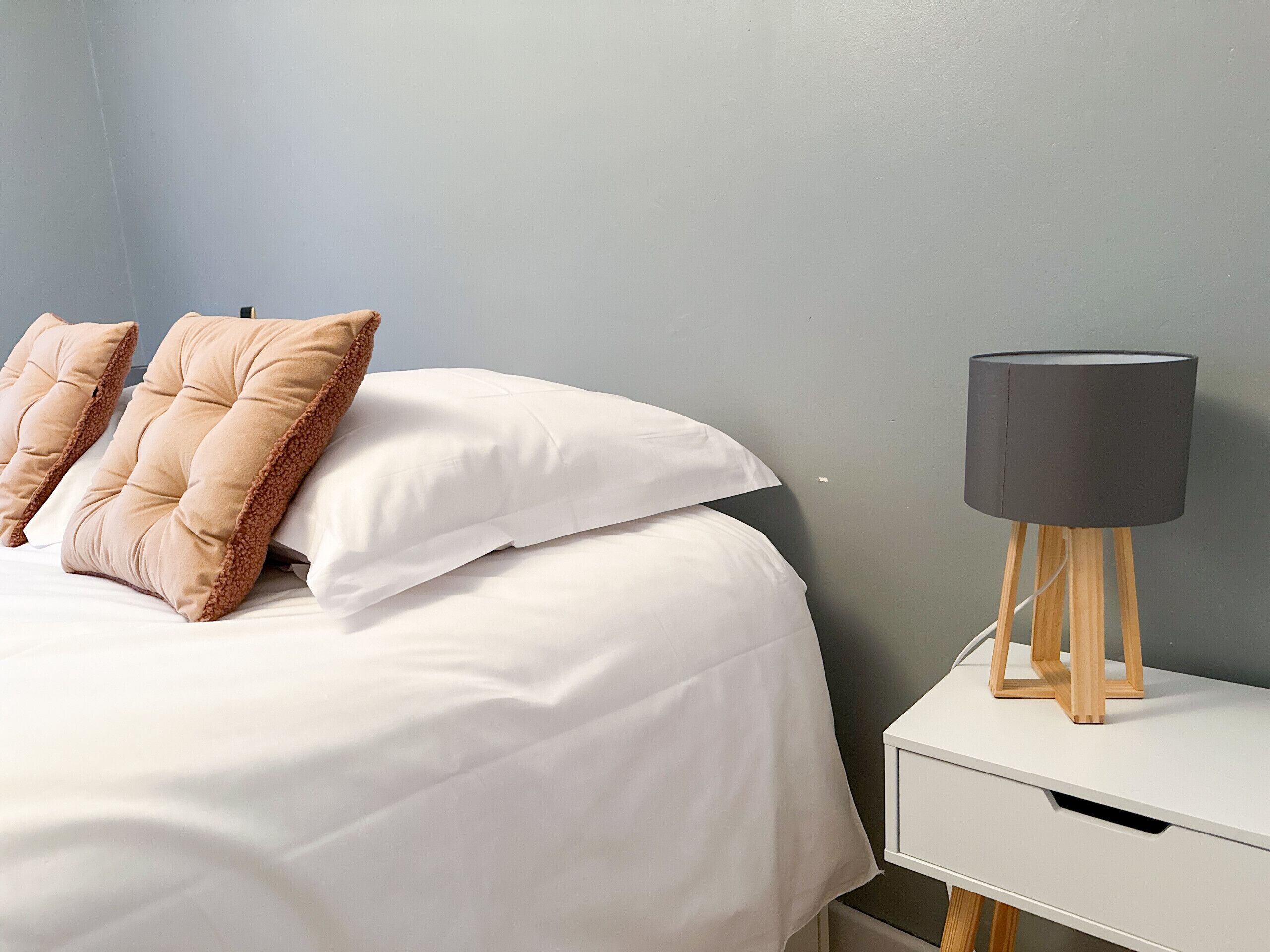 A neatly made bed with white and beige pillows, a white bedside table with a lamp, and a grey wall.