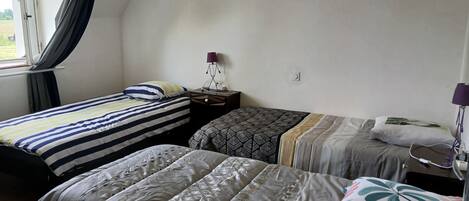 A room with two beds, a window with curtains, a bedside table with a lamp, and a wall socket.