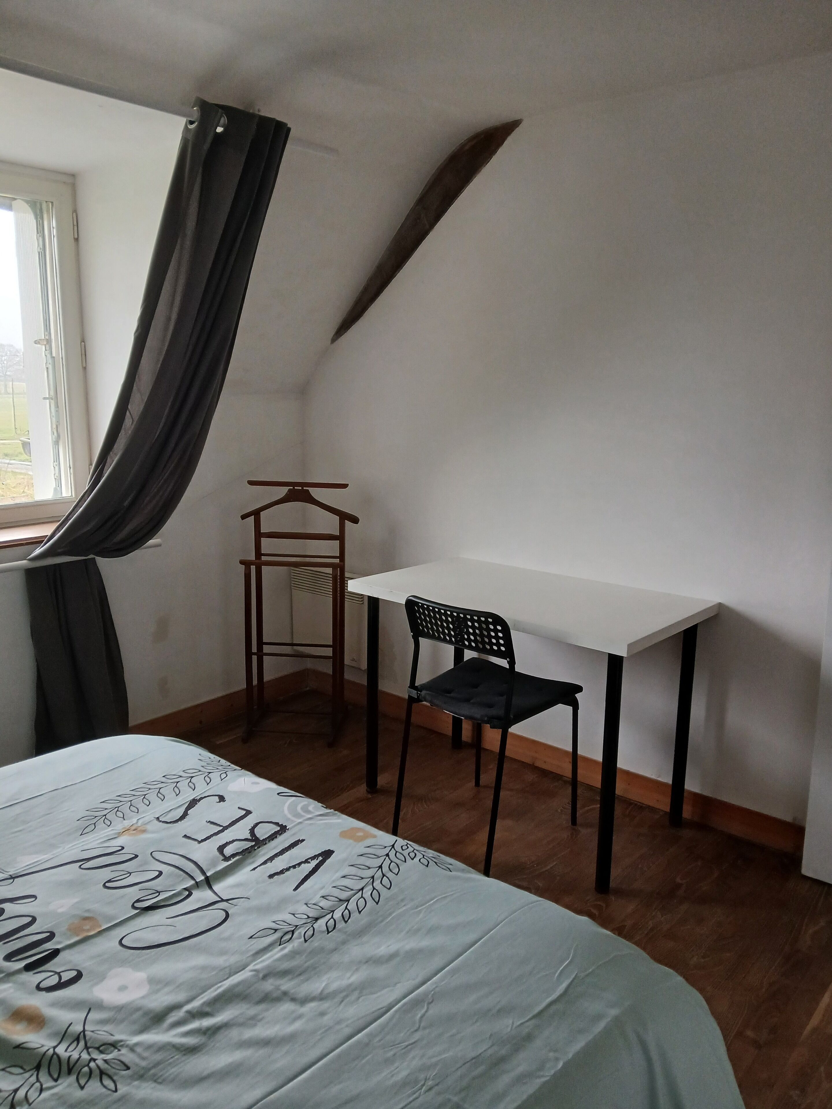 Une petite pièce comprenant un lit, un bureau et une chaise. On y trouve une fenêtre avec des rideaux, un radiateur et un plancher en bois.