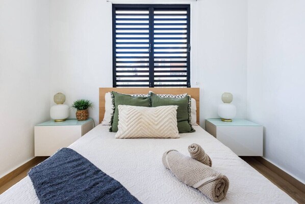 Una cama bien hecha con una colcha blanca y azul a rayas, dos almohadas y una toalla enrollada. Hay dos mesitas de noche con lámparas y una pequeña planta en cada una. Una ventana con persianas horizontales está sobre la cama.