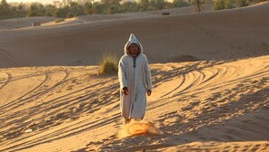 A person walking in a desert with sand dunes and sparse vegetation. - Merzouga Traditional Camp (Taouz)