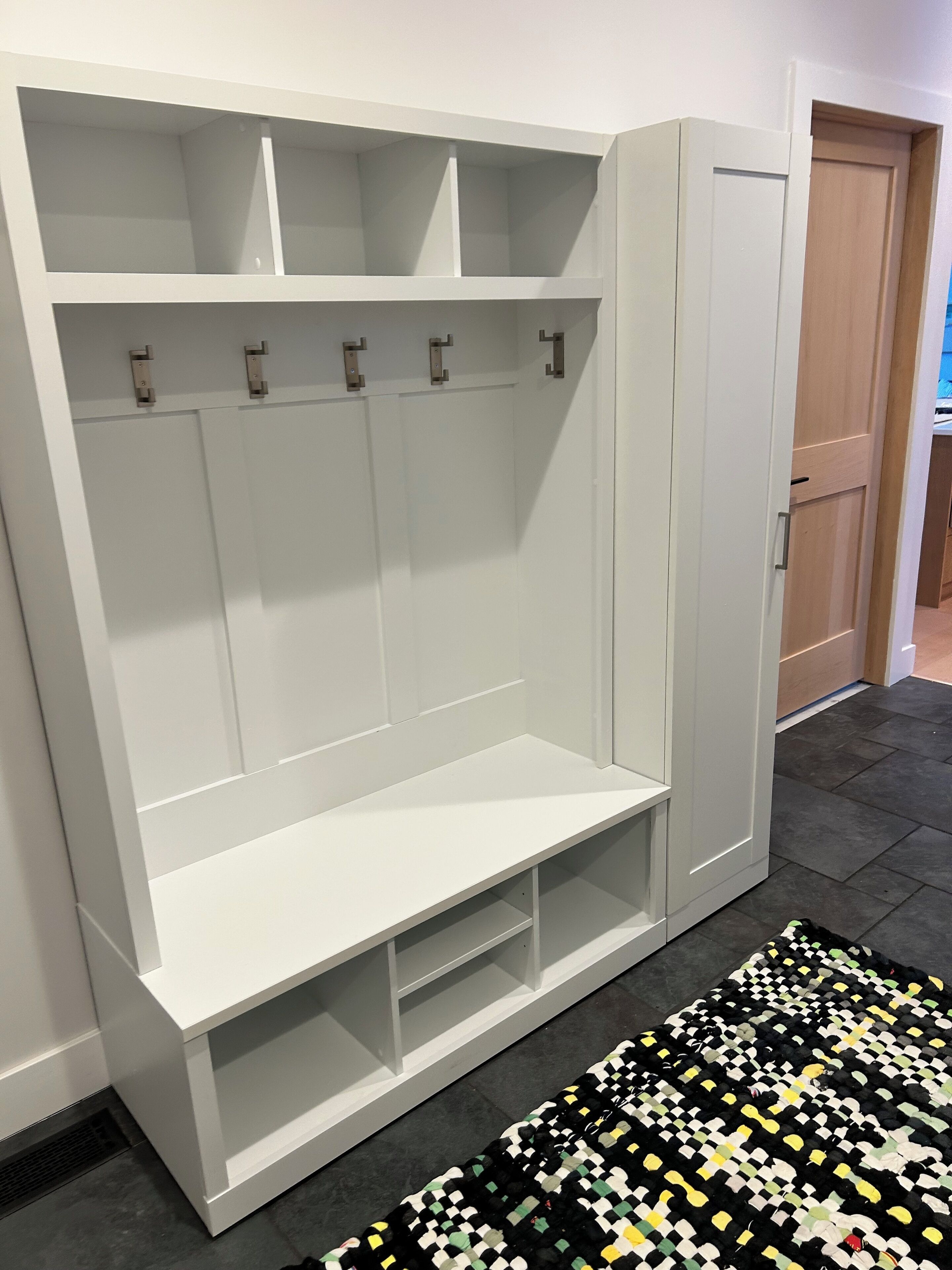 A white wardrobe with multiple shelves and hanging space, a bench at the bottom, and a patterned rug on the floor.