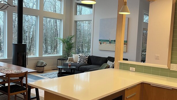 Une cuisine moderne dotée d’un grand îlot central, de meubles en bois et d’un four encastré. On y trouve un coin repas avec une table et des chaises, ainsi qu’un canapé à l’arrière-plan.