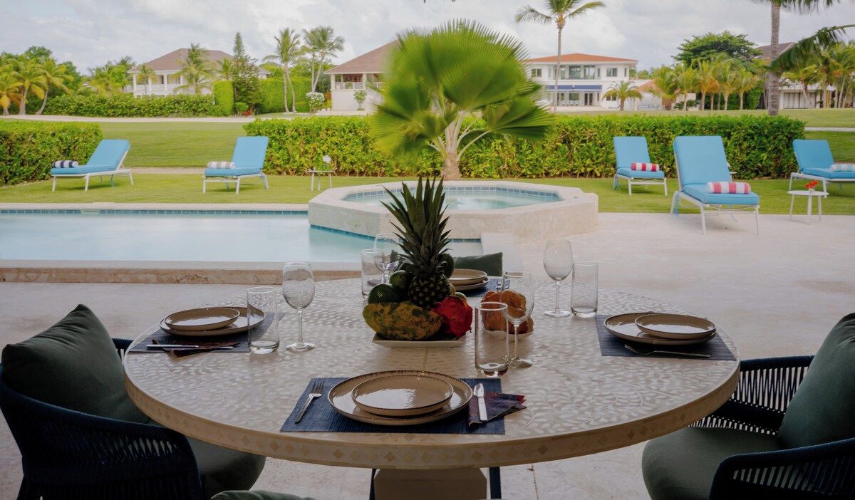 Une table ronde dressée pour un repas, agrémentée d’une décoration tropicale au centre, entourée de fauteuils confortables et avec une piscine en arrière-plan.