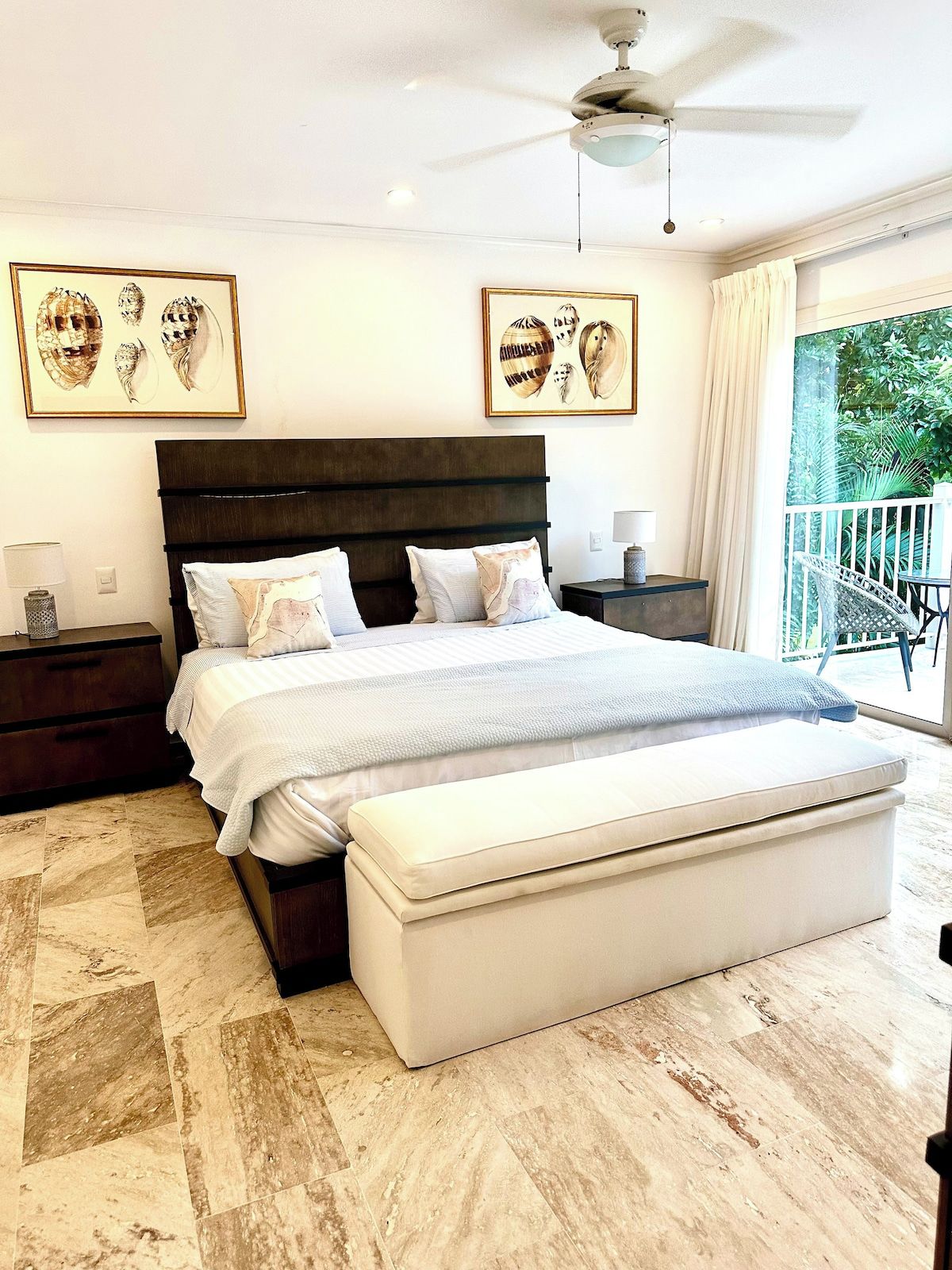A bedroom with a large bed, a ceiling fan, artwork on the walls, and a balcony with a view of greenery.