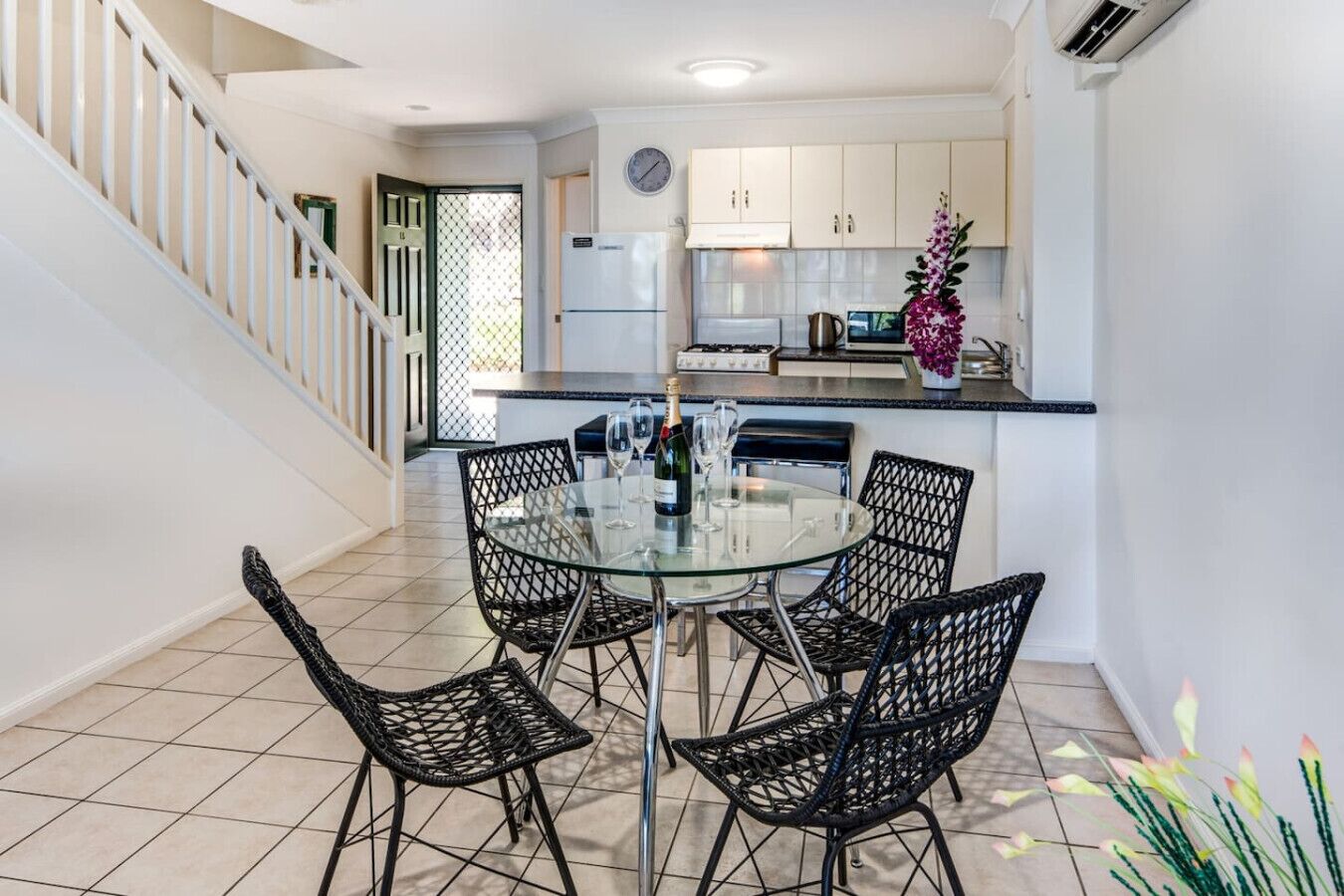 A modern kitchen with white cabinets, a stainless steel refrigerator, and a dining area with a glass table and wicker chairs.