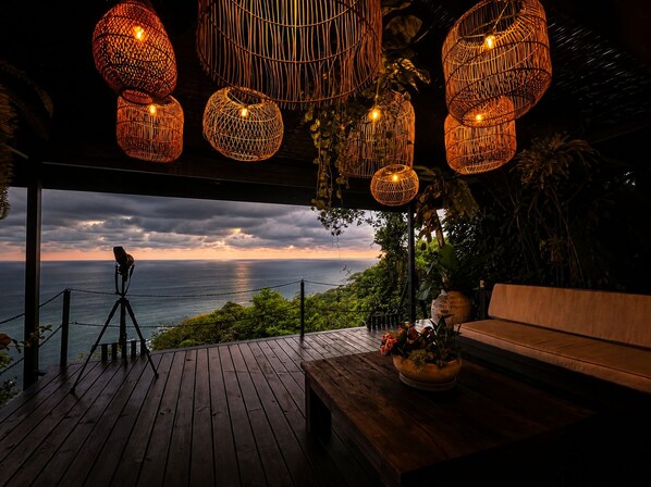 Una terraza de madera con un banco y un telescopio, con vistas a la costa y un atardecer.