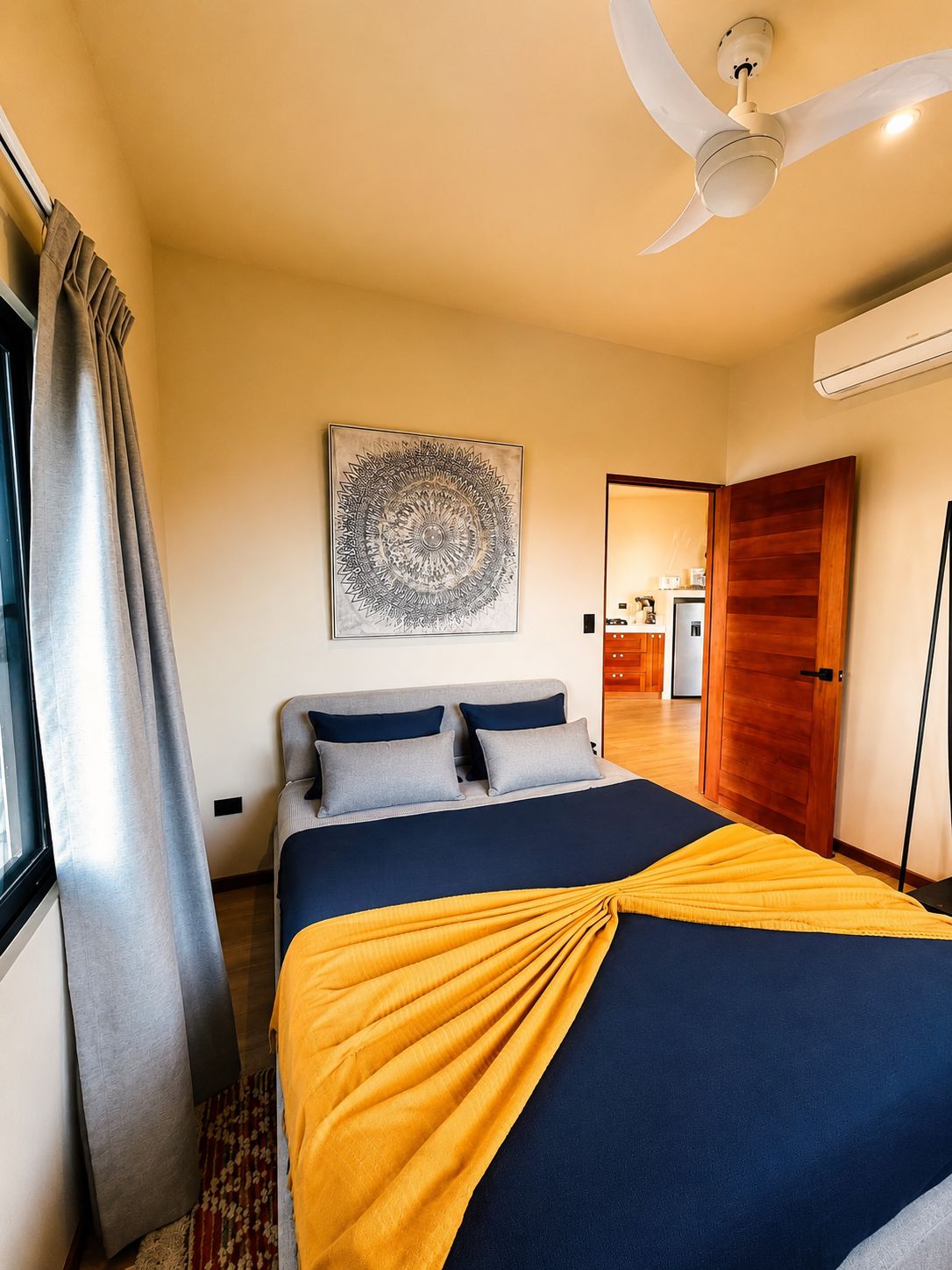 Un dormitorio con cama, ventilador de techo, aire acondicionado de pared y una puerta que da a una cocina.