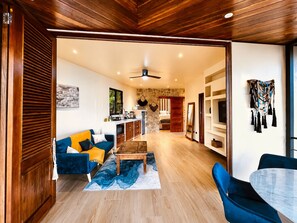 Un vaste espace de vie comprenant un canapé, une table basse et une table à manger. On aperçoit une cuisine au fond et un mur en pierre.