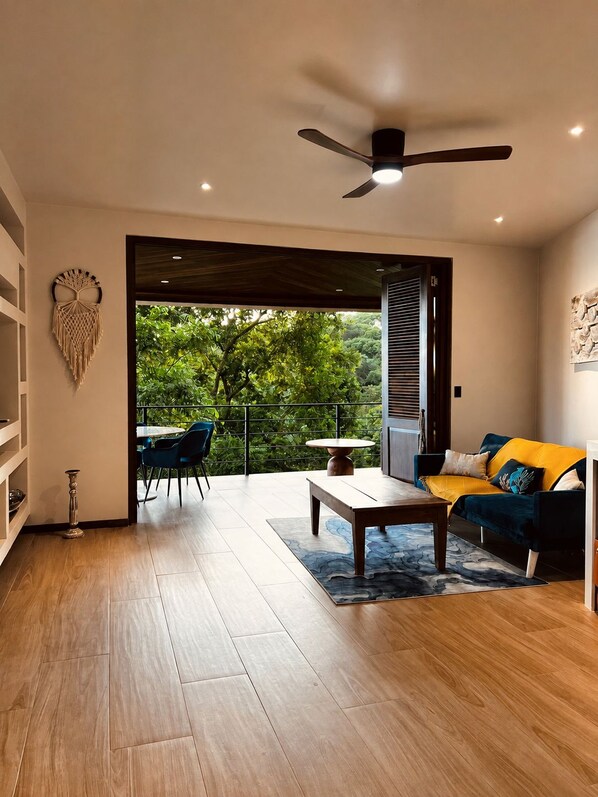 A modern living room with a wooden floor, a ceiling fan, a sofa, a coffee table, and a view of greenery outside.