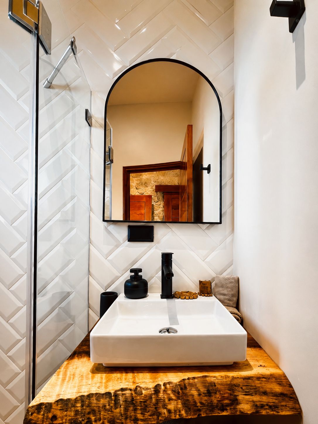Un baño con pared de azulejos blancos, tocador de madera, lavamanos blanco, grifo negro y espejo con arco.