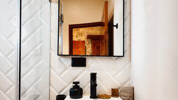 Un baño con pared de azulejos blancos, tocador de madera, lavamanos blanco, grifo negro y espejo con arco.