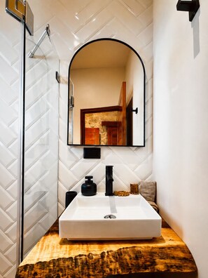Une salle de bain avec un mur recouvert de carreaux blancs, un meuble-lavabo en bois, un lavabo blanc, un robinet noir et un miroir surmonté d’un arc.