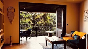 Een moderne woonkamer met houten vloer, een plafondventilator, een bank, een salontafel en uitzicht op groen buiten.