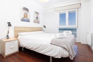 A neatly arranged bedroom with a wooden headboard, a white bed, a nightstand with a lamp, and a window offering a view of the sea.