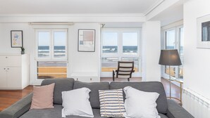 Ein modernes Wohnzimmer mit grauem Sofa, weißen und gestreiften Kissen, Holzboden und großen Fenstern mit Blick auf den Strand.