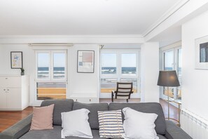 A modern living room with a grey sofa, white and striped pillows, a wooden floor, and large windows offering a view of the beach.