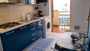 A compact kitchen with a gas stove, sink, and blue cabinets. There is a dining table with a floral tablecloth and a chair. A balcony with a pool is visible through the open door. - Apartment 'Balcone Sul Golfo' with Sea View, Wi-Fi and Air Conditioning (Alcamo)