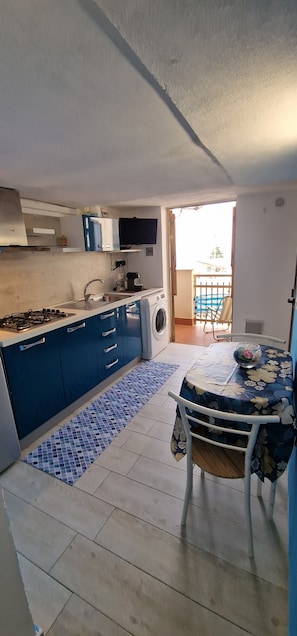 Cocina compacta con estufa a gas, pileta y muebles azules. Se observa una mesa de comedor con mantel floral y una silla. Se ve un balcón con piscina a través de la puerta abierta.