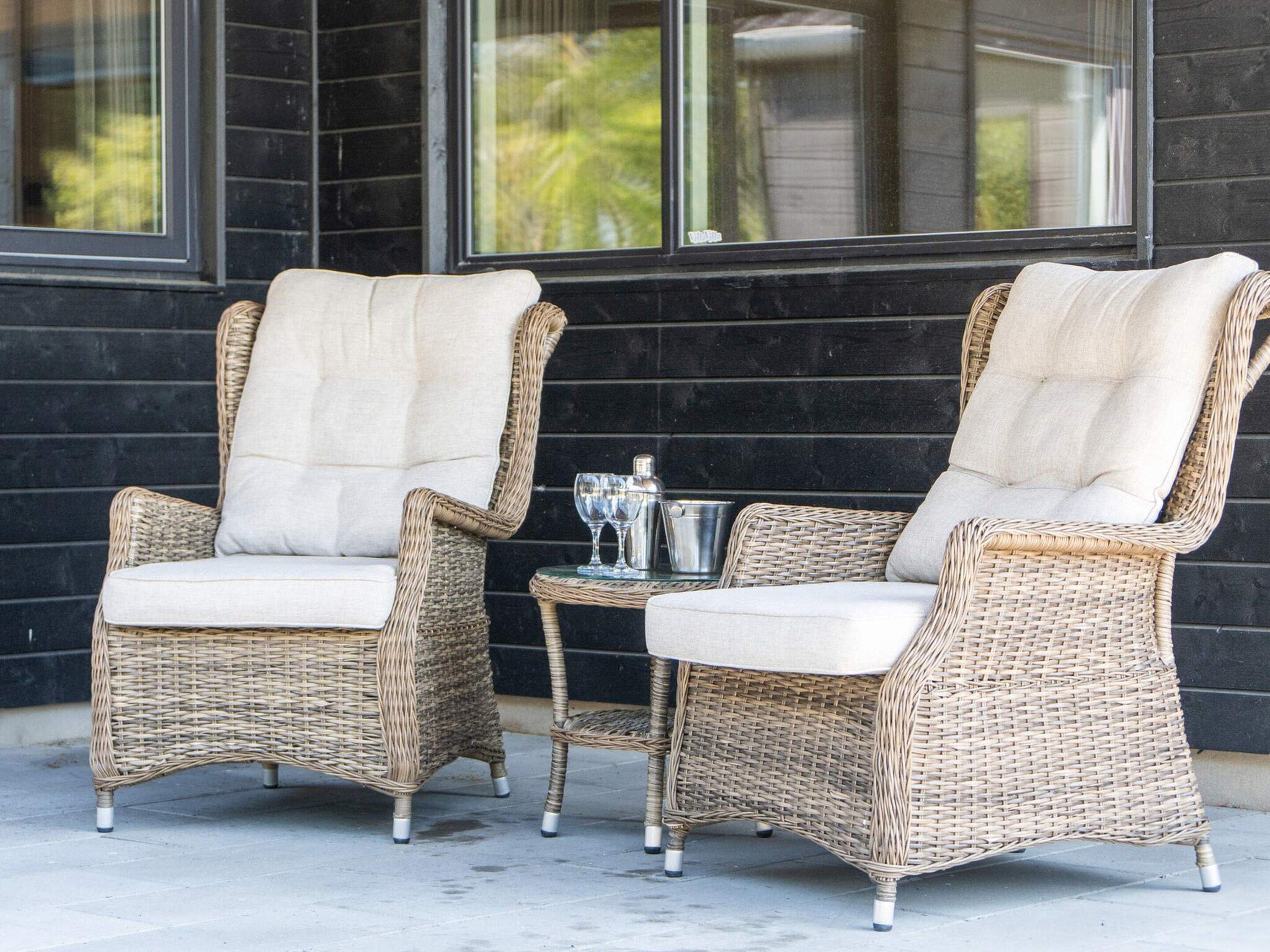 Two wicker chairs with cushions, a metal pot, and a glass on a small table, positioned outside a building with large windows.