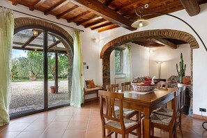 An open-plan living area with a dining table, chairs, and a sofa. There are arched brick walls and a ceiling with exposed beams.