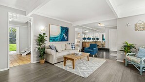 A spacious living room with a sofa, coffee table, and chairs, a dining area with a kitchen in the background, and a large painting on the wall.
