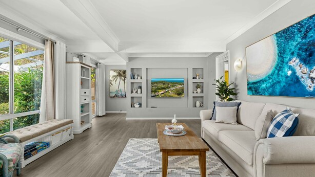 A spacious living room with a large window, a wooden coffee table, a beige sofa, and a bookshelf.