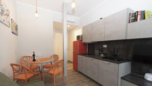 Et kompakt køkken med lysegrå skabe, et rødt køleskab og et spiseområde med orange stole og et bord.