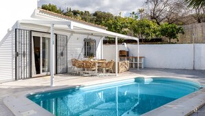 Ein Schwimmbad mit Terrassenbereich, Grill und überdachter Sitzgruppe.
