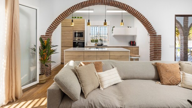 Ein modernes Wohnzimmer mit einer Couch, einem Essbereich und einer dahinterliegenden Küche, sowie einem großen Fenster.