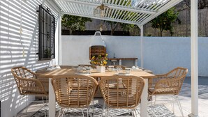 Eine überdachte Terrasse mit Esstisch für vier Personen, umgeben von Korbstühlen, einem Grill und einer hängenden Lampe.