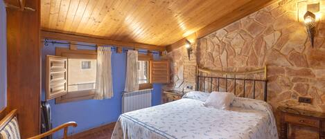 Un dormitorio con techo de madera, pared de piedra, cama con estructura metálica, una silla de madera y una mesita de noche.