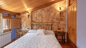 A bedroom with a stone wall, wooden ceiling, a bed with patterned bedding, and a nightstand with a lamp.