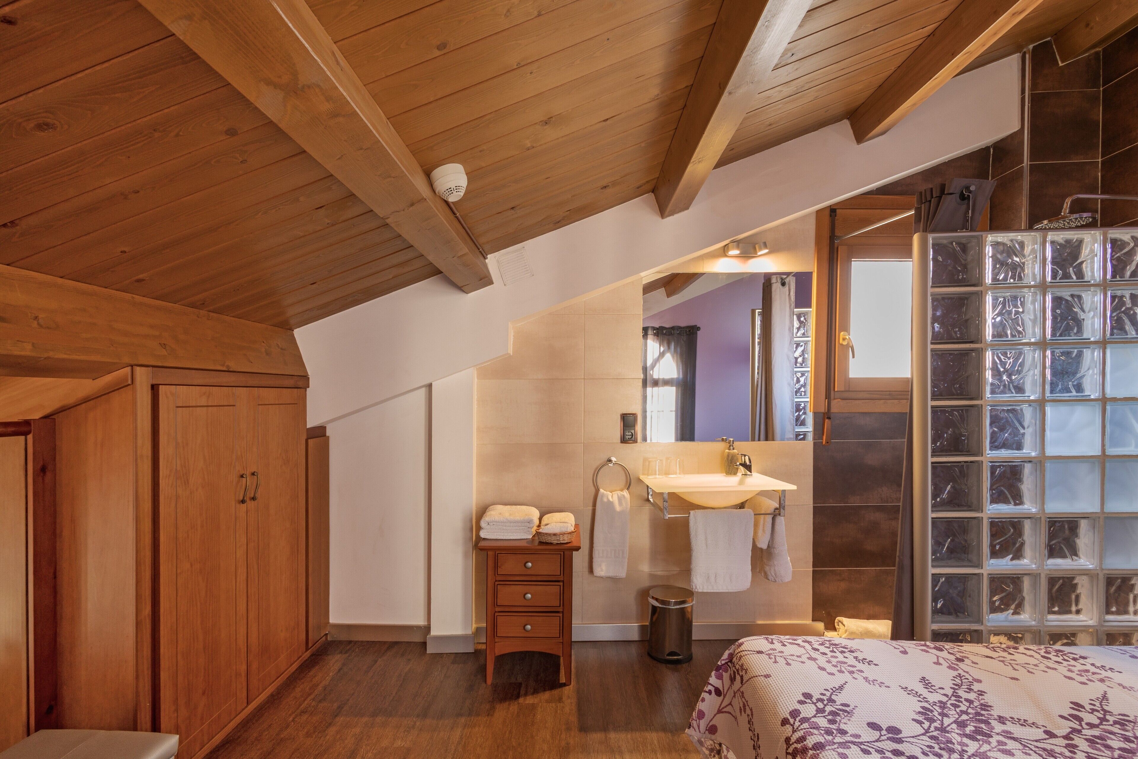 Un dormitorio con vigas de madera en el techo, un armario de madera, una mesita de noche, una cama con colcha estampada, y un baño con lavabo, espejo y cabina de ducha con bloques de vidrio.