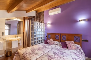 Un dormitorio con techo de madera, pared morada, cama con ropa de cama floral, baño con lavamanos y espejo, y ventana con vidrio esmerilado.